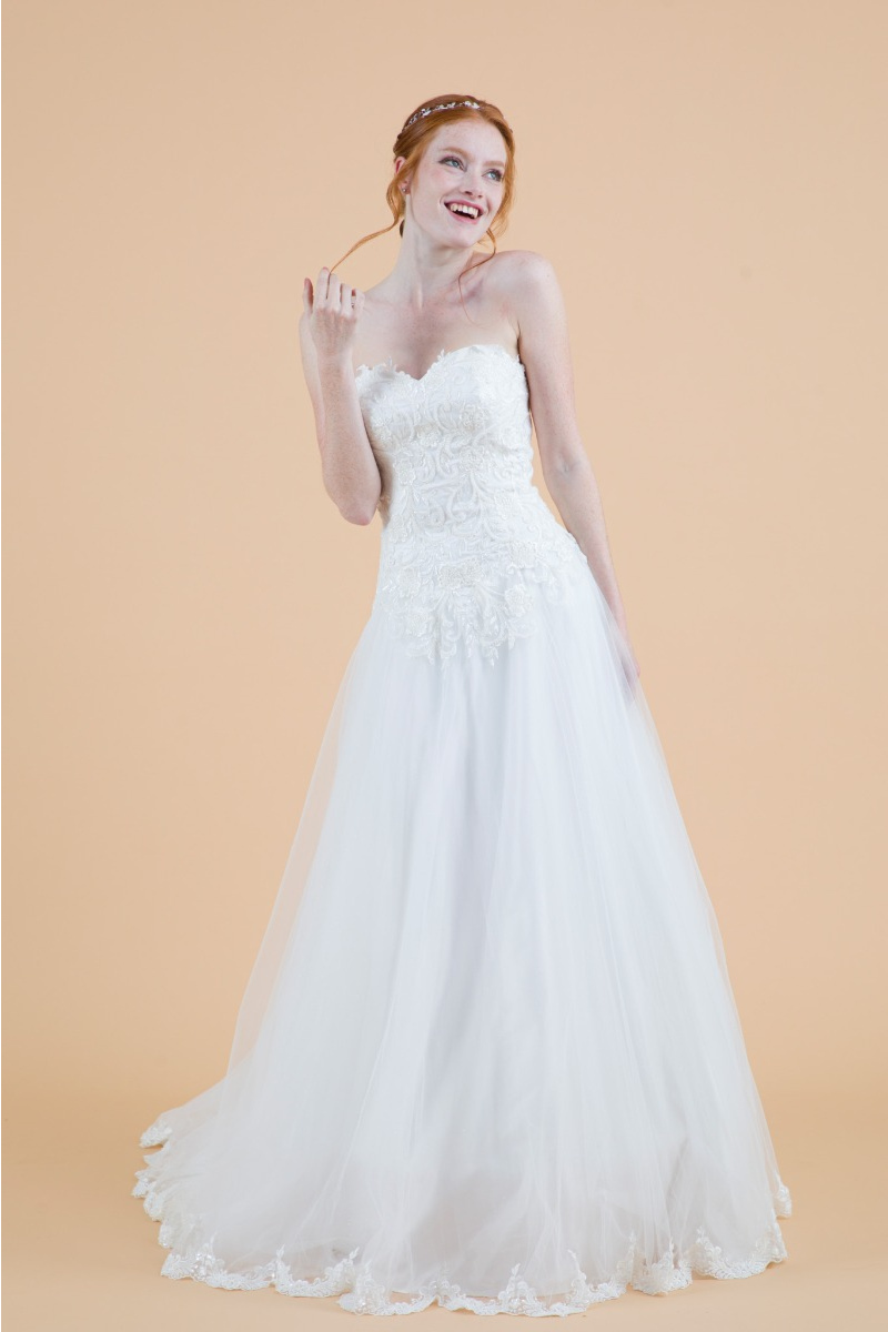 Abito da sposa Camomilla in pizzo bianco con corpetto ricamato e gonna ampia, sfondo beige.
