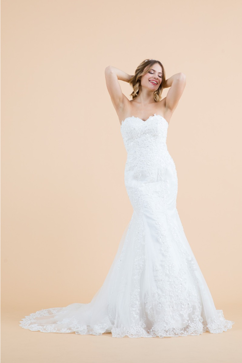 Abito da sposa geranio bianco, senza spalline, con dettagli in pizzo e gonna svasata su sfondo beige.