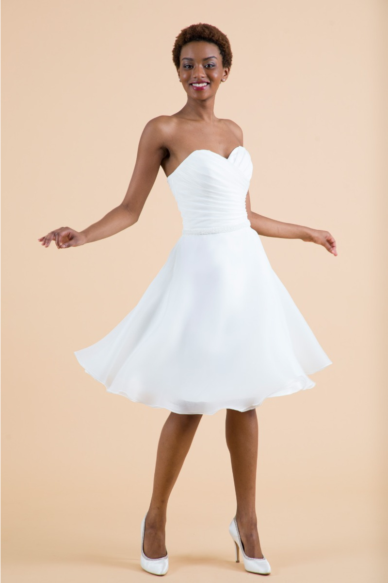 Abito da sposa Cisto corto, senza spalline, con gonna ampia e scarpe col tacco, sfondo neutro.