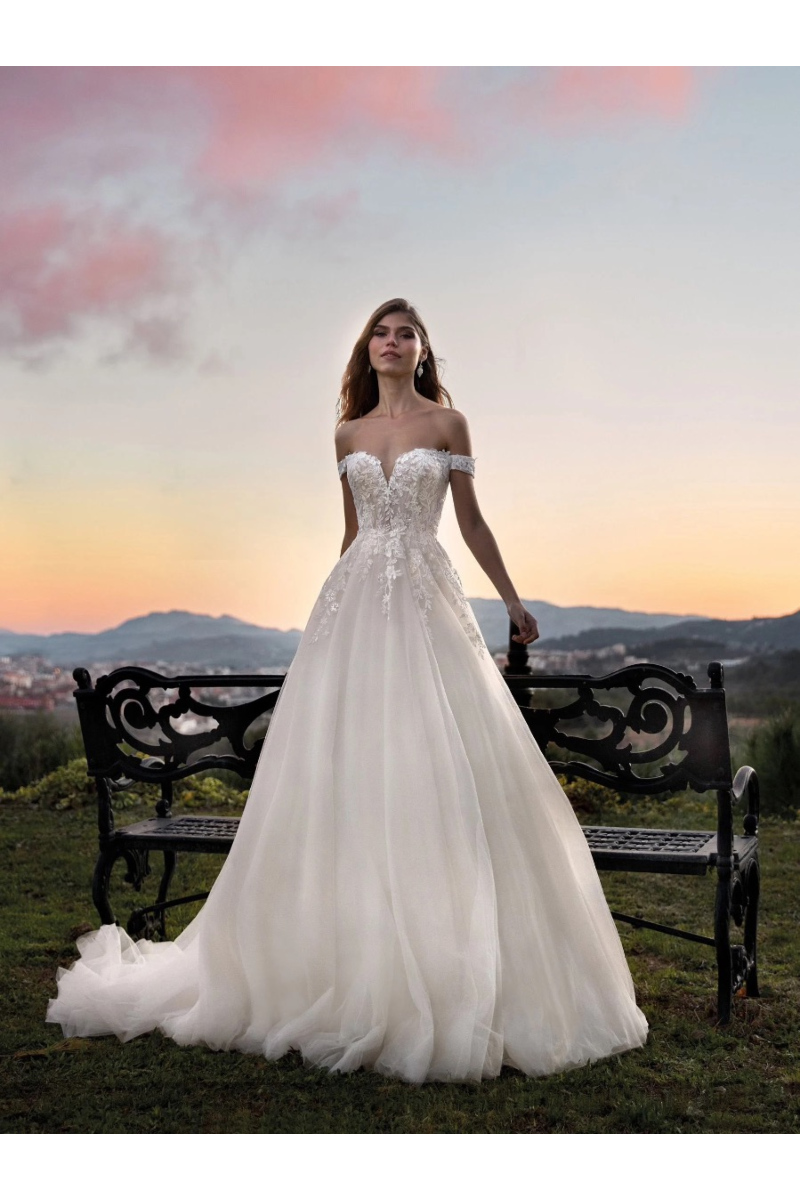 Abito da sposa KAEDA Nicole Colet, elegante e romantico, con sfondo tramonto e panchina decorativa.
