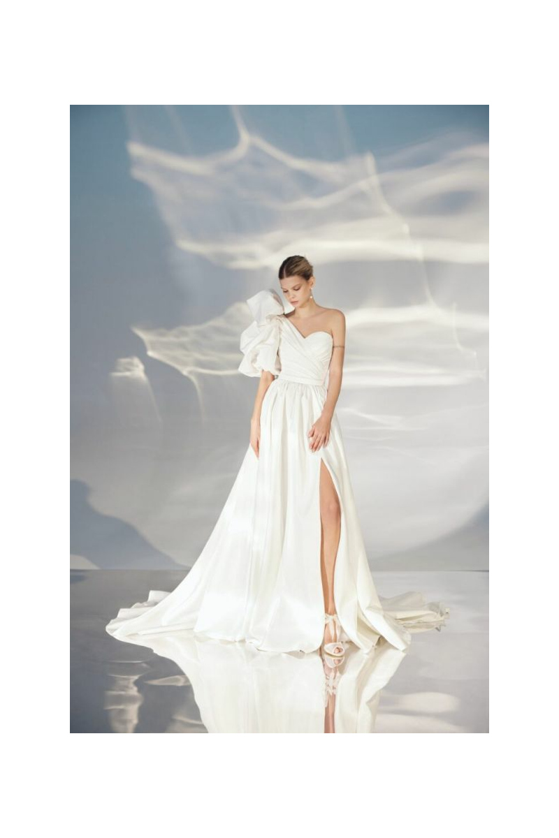 Abito da sposa bianco L317 ARIAMO LIGHT con spacco e manica asimmetrica, su sfondo etereo.