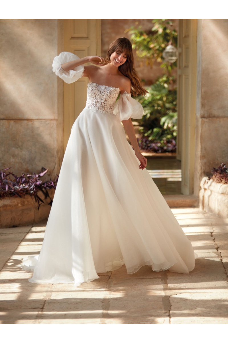 Abito da sposa NASRIN con corpetto in pizzo e maniche voluminose, ambientato in un giardino soleggiato.