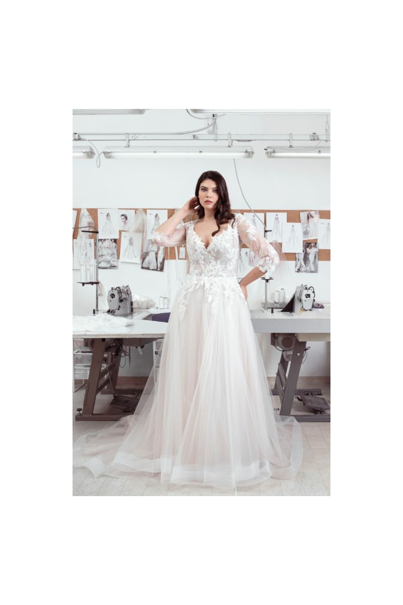 Abito da sposa curvy Adrienne in pizzo e tulle, maniche lunghe, atelier sartoriale sullo sfondo.
