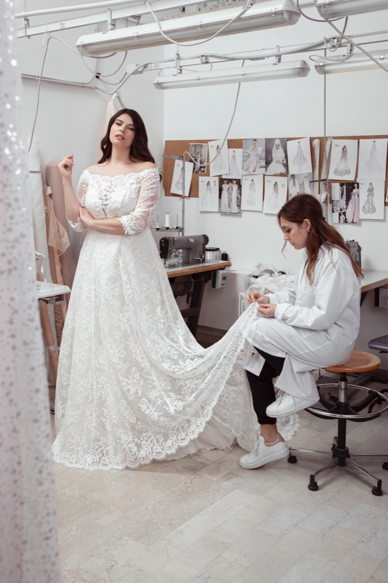 Sposa curvy in abito bianco di pizzo con sarta al lavoro in atelier di moda.