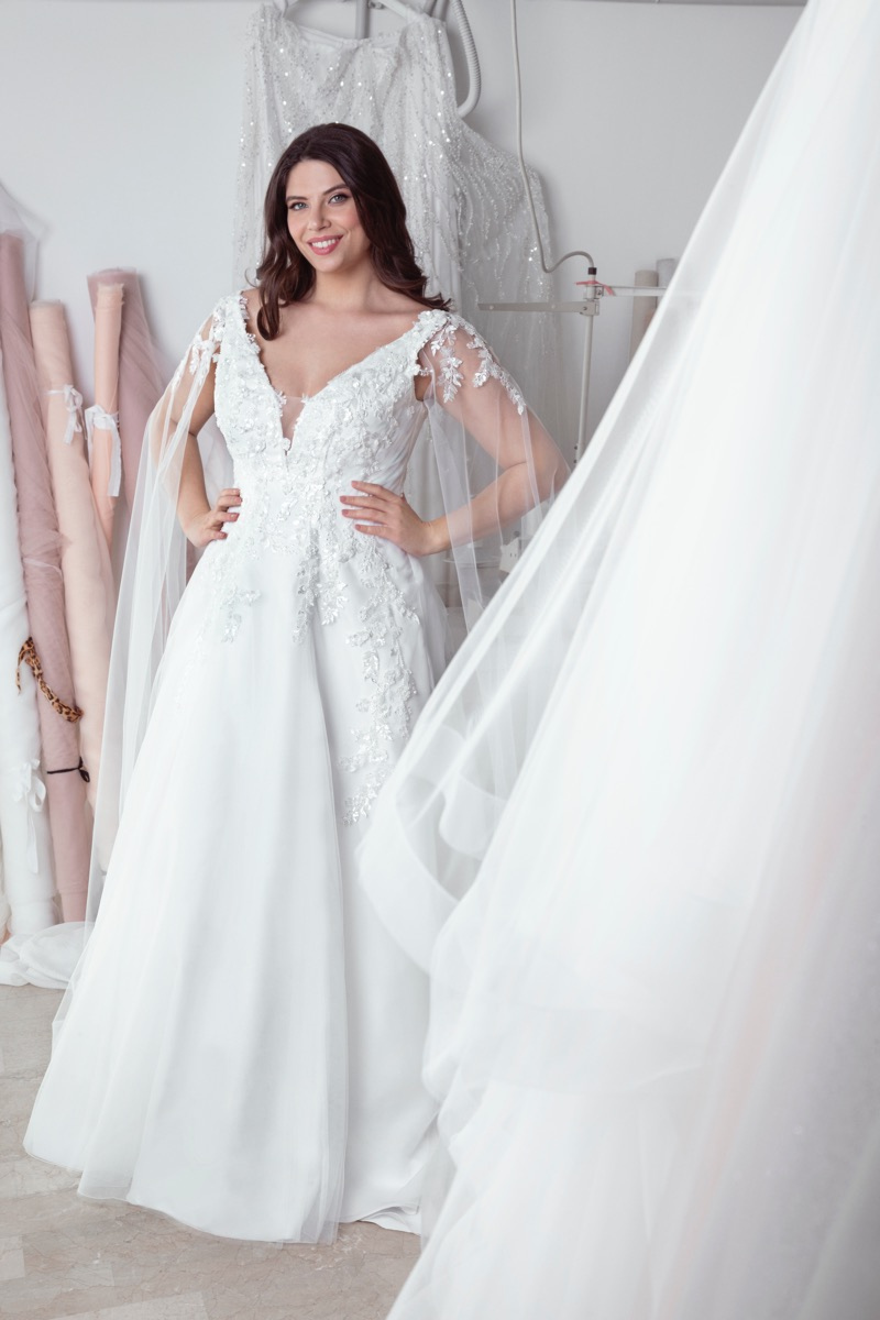 Abito da sposa curvy Freya, elegante e ricamato, indossato da modella sorridente in atelier di moda.