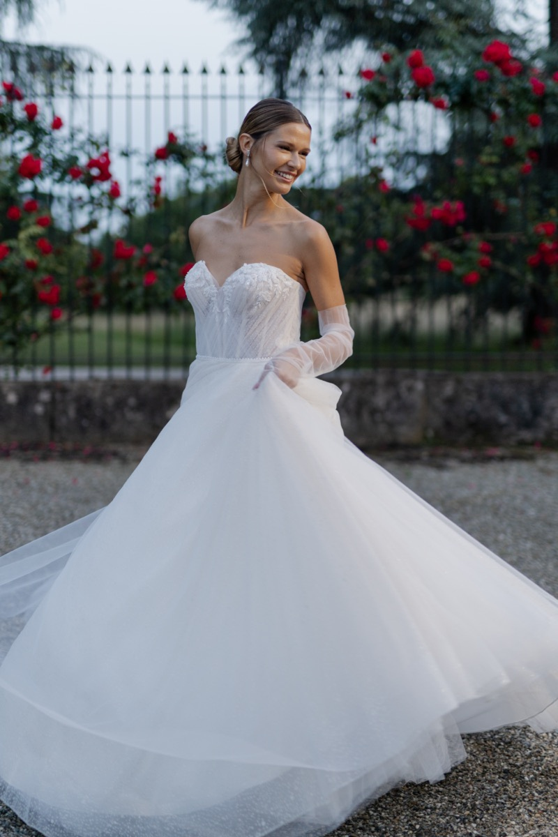 Abito da sposa JACLYN di SPOSAE, design romantico con corpetto in pizzo, sfondo floreale.