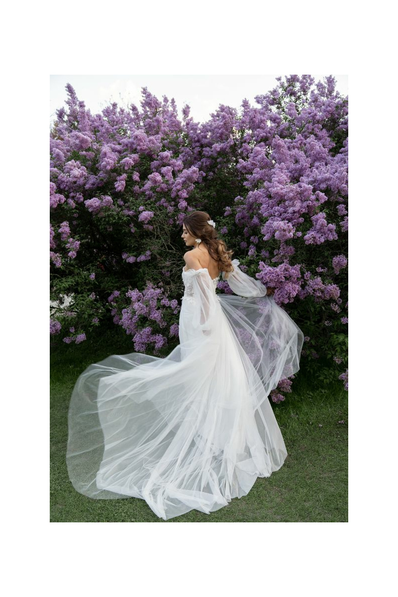 Abito da sposa bianco con velo lungo, donna di spalle tra cespugli di fiori viola in giardino.