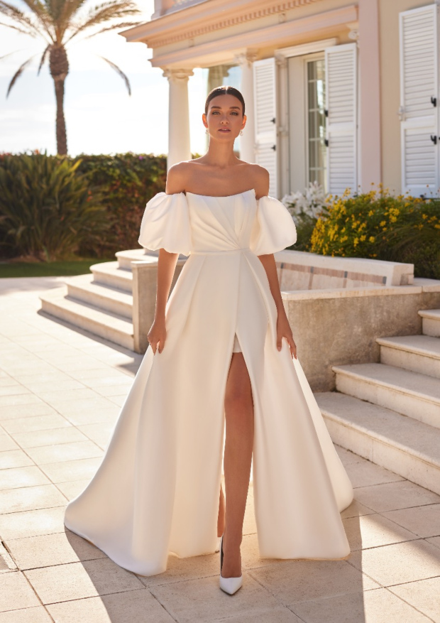 Abito da sposa ANGLESEY con spalle scoperte e spacco, ambientato in un elegante giardino con palme.