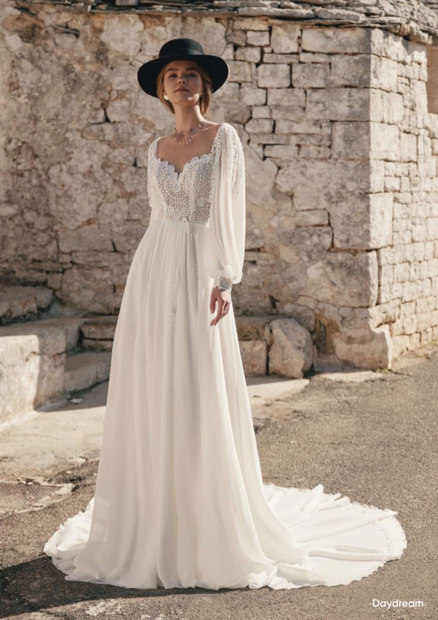 Abito da sposa boho DAYDREAM ARIAMO con maniche lunghe e cappello nero, sfondo rustico in pietra.