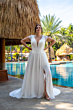Abito da sposa Bella by Amelie 30113W, scollo a V, pizzo, sfondo tropicale, piscina, atmosfera elegante.
