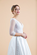 Abito da sposa Silene in pizzo bianco con maniche trasparenti e gonna ampia, sfondo beige.